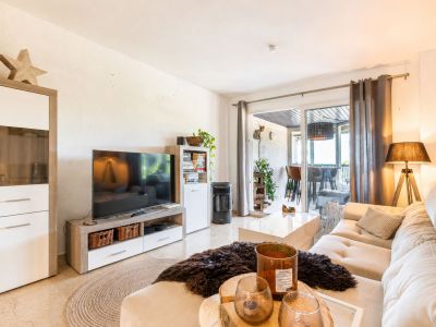 Salon lumineux avec un canapé d'angle clair, une table basse blanche brillante et un meuble TV. Des portes coulissantes en verre s'ouvrent sur une terrasse couverte.