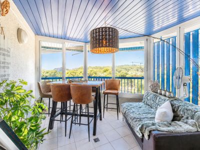 Terrasse vitrée avec plafond à lattes bleues, offrant des vues panoramiques sur la verdure. Elle intègre un coin repas informel avec tabourets hauts et un espace salon confortable.