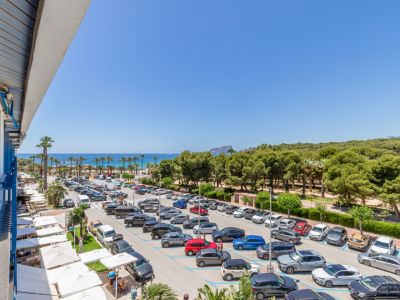 Vue panoramique depuis la terrasse, offrant la mer Méditerranée, une plage bordée de palmiers et le Peñón de Ifach. En contrebas, un parking et des auvents blancs.