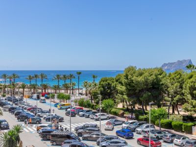 Vue panoramique sur la mer Méditerranée, avec des palmiers, une plage de sable et le Peñón de Ifach au loin, bordée par un parking animé et des pins.