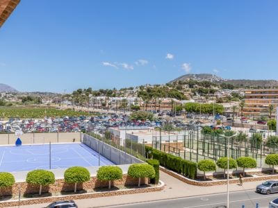 Vue panoramique sur un terrain de sport bleu, plusieurs courts de padel, un vaste parking et le paysage urbain de Moraira avec des montagnes lointaines.
