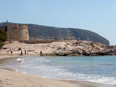 Plage de sable avec une tour de guet historique en pierre sur un promontoire rocheux, bordée par les eaux calmes de la Méditerranée sous un ciel clair.