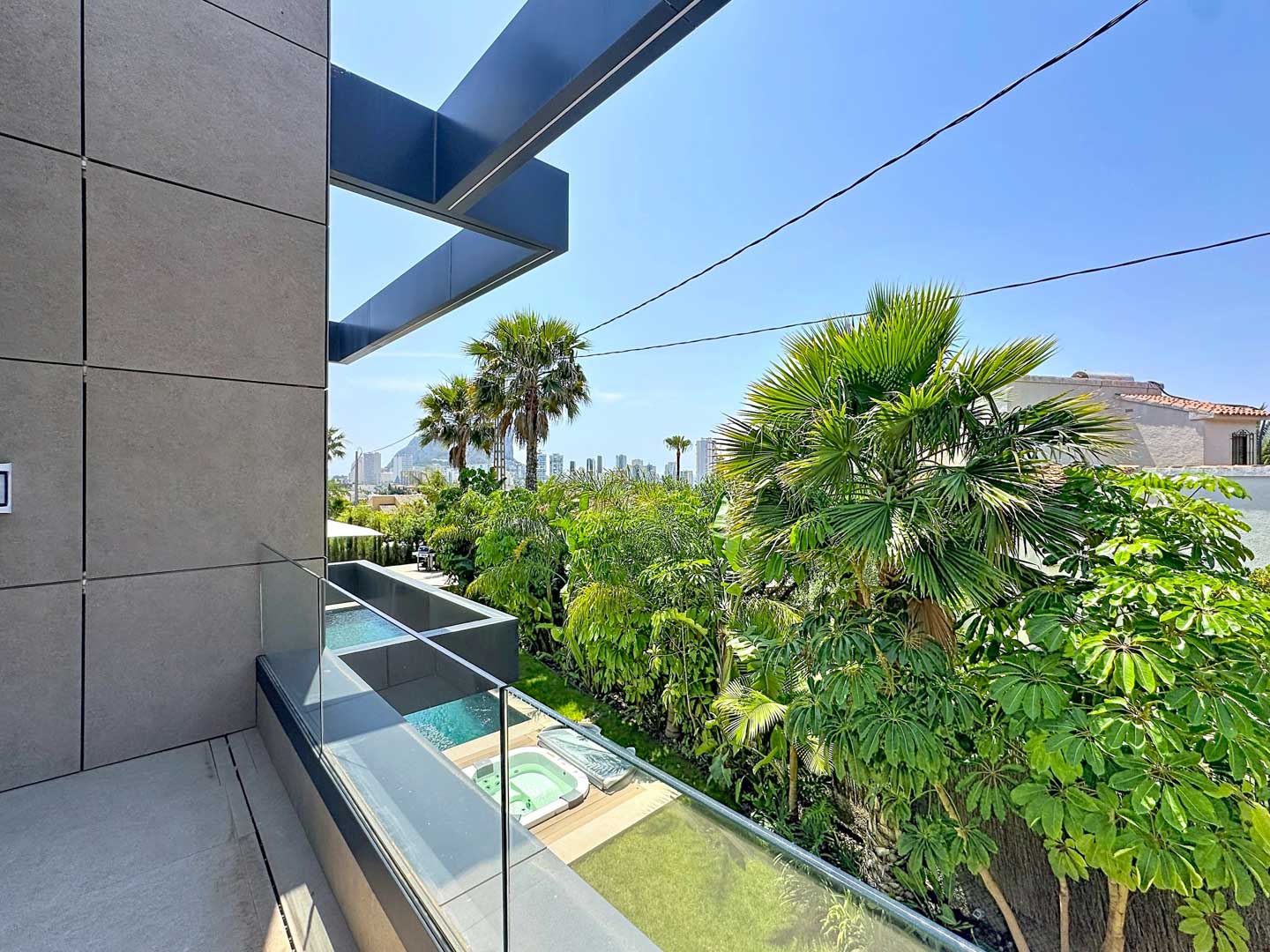 Balcon moderne avec façade à panneaux gris et garde-corps en verre, dévoilant des jacuzzis extérieurs, une végétation tropicale luxuriante et la skyline de Calpe.