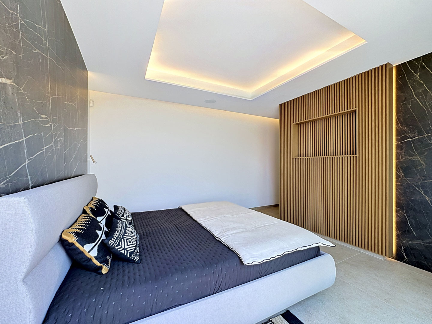 Chambre moderne avec lit tapissé gris clair, panneaux muraux effet marbre foncé et un mur central à lattes de bois avec éclairage intégré et étagère encastrée. Le plafond intègre un éclairage périmétrique.