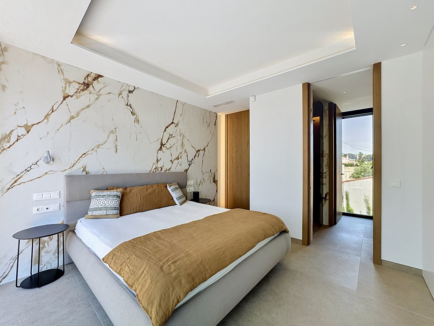 Chambre principale avec tête de lit en tissu et mur d'accent en carreaux effet marbre aux veines dorées. Éclairage encastré au plafond et accès à une zone dressing/salle de bain.