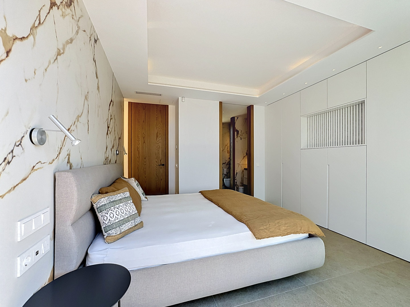 Chambre principale avec un mur d'accent en marbre, un lit tapissé et des armoires blanches intégrées. Une porte en bois mène à la salle de bain attenante.