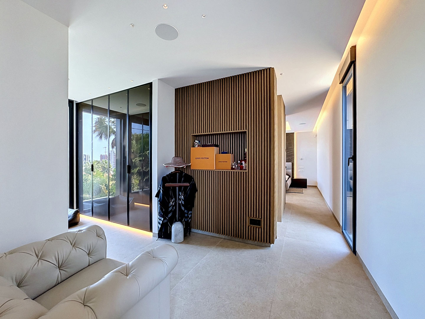 Couloir moderne avec mur d'accent en bois à lattes et niche. De larges baies vitrées s'ouvrent sur une terrasse extérieure verdoyante, éclairé par un éclairage intégré et en corniche.