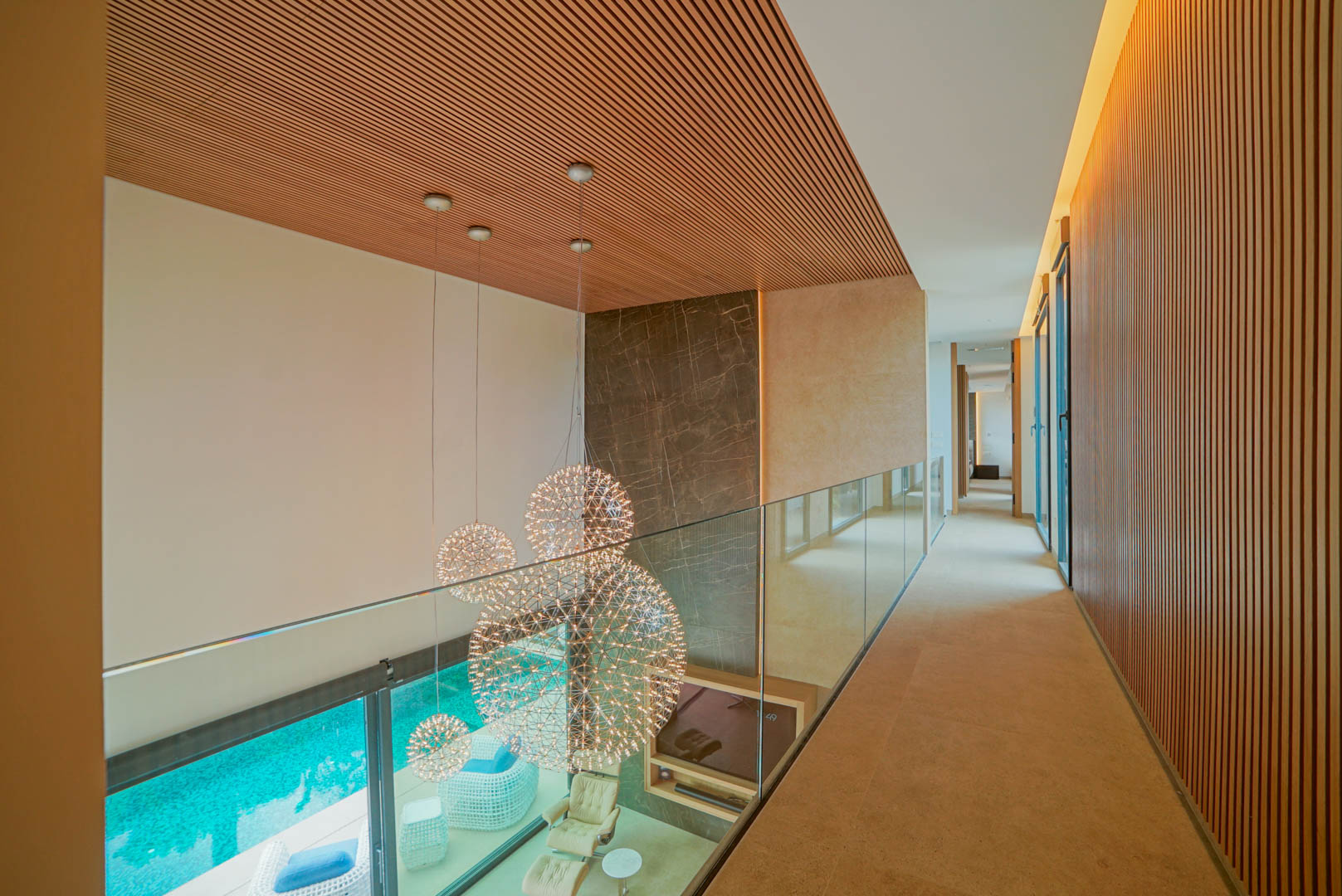 Couloir supérieur avec garde-corps en verre et plafond à lattes de bois, surplombant un salon double hauteur avec lustres sphériques et piscine.