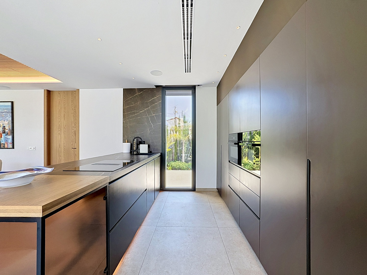 Cuisine design avec meubles sombres sans poignées et électroménager Miele intégré. L'îlot central présente un plan de travail noir, une plaque et un évier, complété par un mur en pierre veinée et un accès jardin.