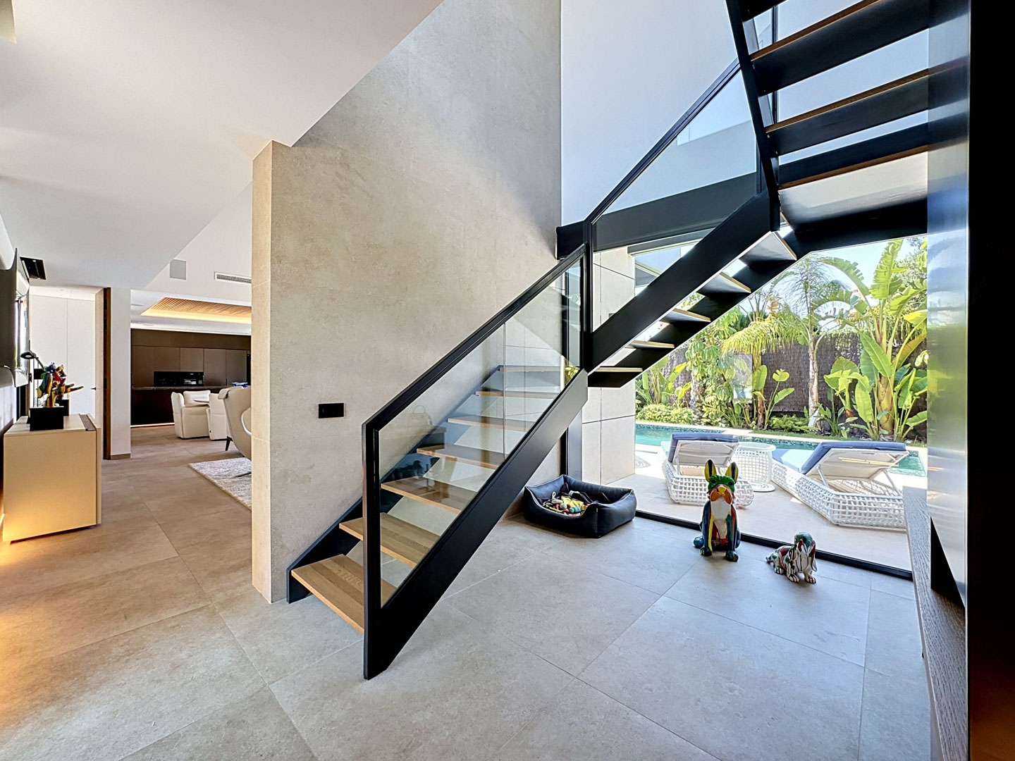 Escalier moderne à marches ouvertes avec garde-corps en verre et métal noir, reliant l'intérieur lumineux et offrant une vue sur la piscine et le jardin tropical.