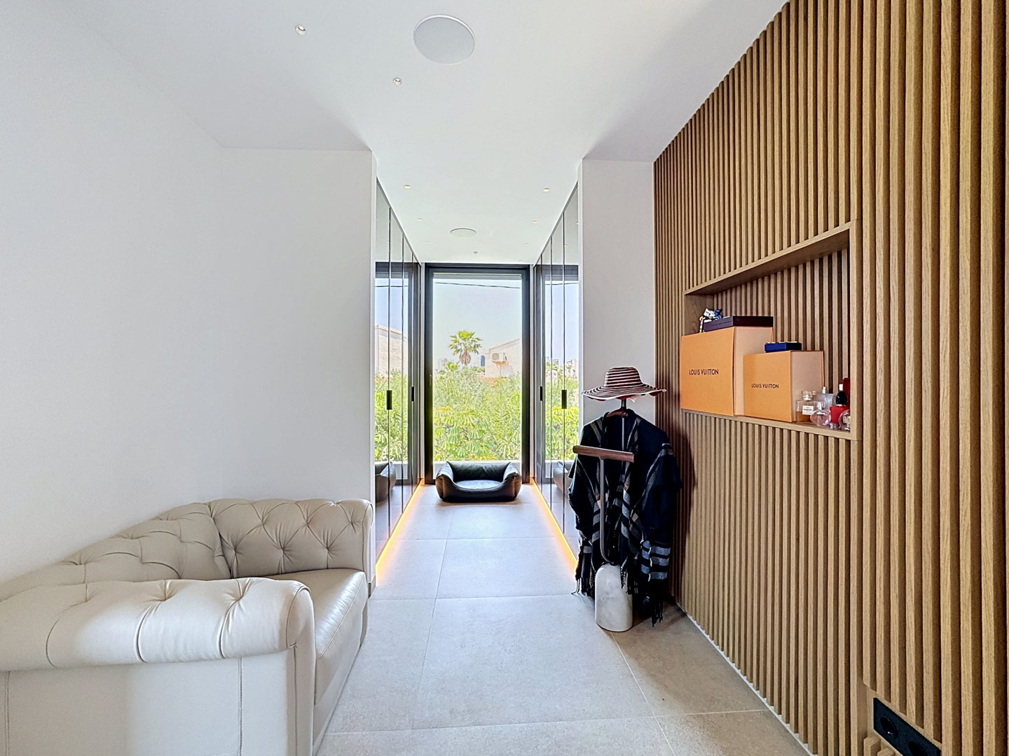 Espace dressing raffiné avec mur à lattes de bois et étagère intégrée, un canapé en cuir capitonné et de larges portes vitrées donnant sur la verdure extérieure.