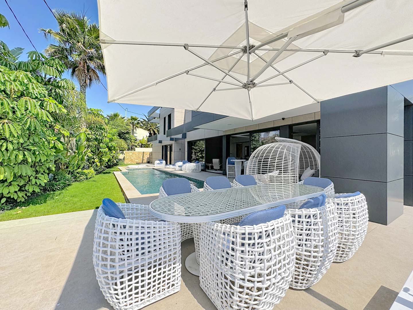 Espace repas extérieur avec table en verre et chaises tressées blanches aux coussins bleus, abrité par un grand parasol. La piscine privée et le jardin tropical bordent la façade moderne de la villa.