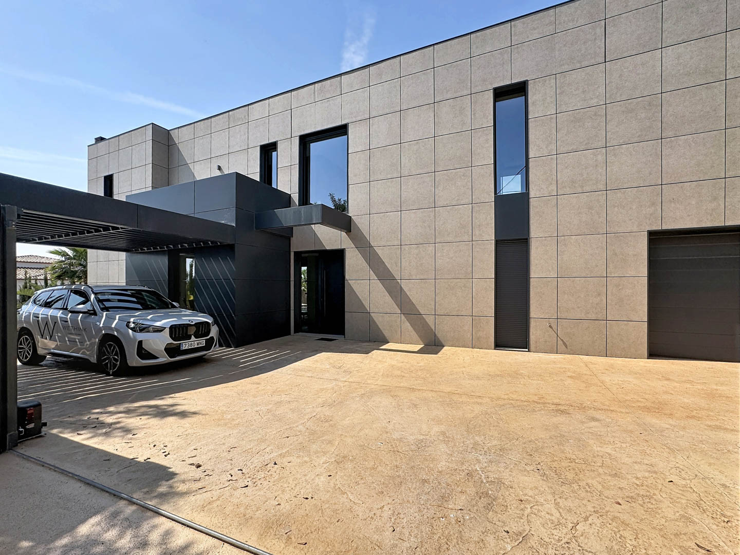 Extérieur contemporain de la villa avec façade en panneaux clairs et entrée anthracite. Un carport à lames protège le véhicule, jouxtant une allée pavée texturée et le garage intégré.