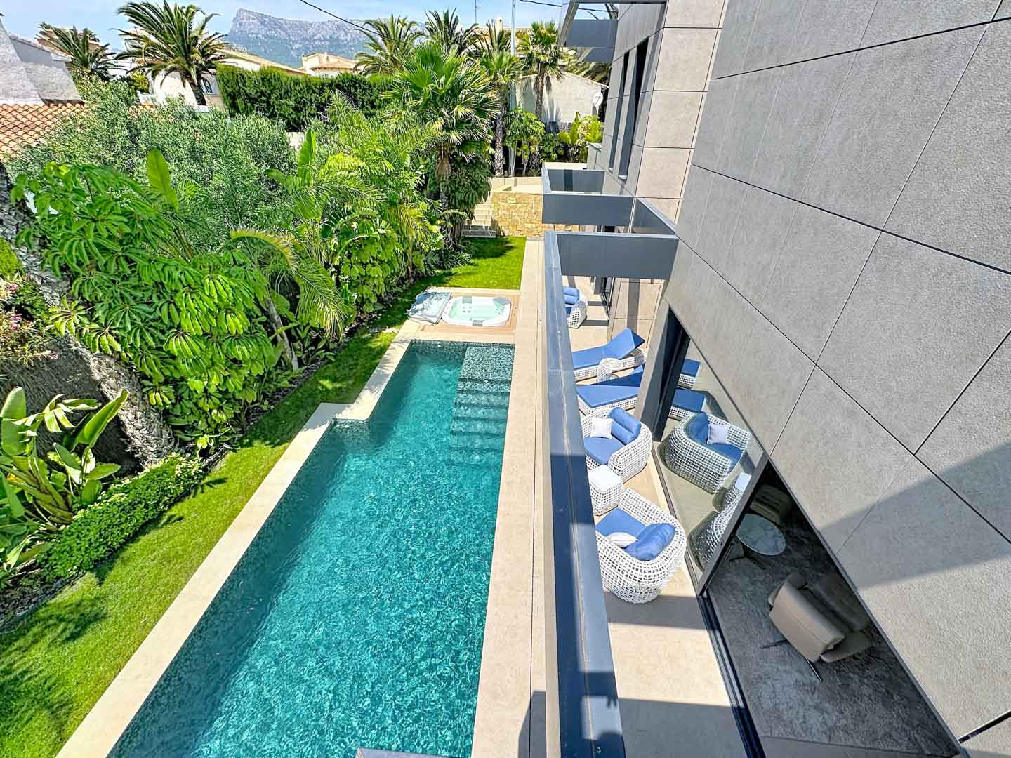 Piscine rectangulaire avec jacuzzi intégré, entourée de jardins tropicaux luxuriants et de pelouse. Balcons modernes meublés et façade en carreaux gris.