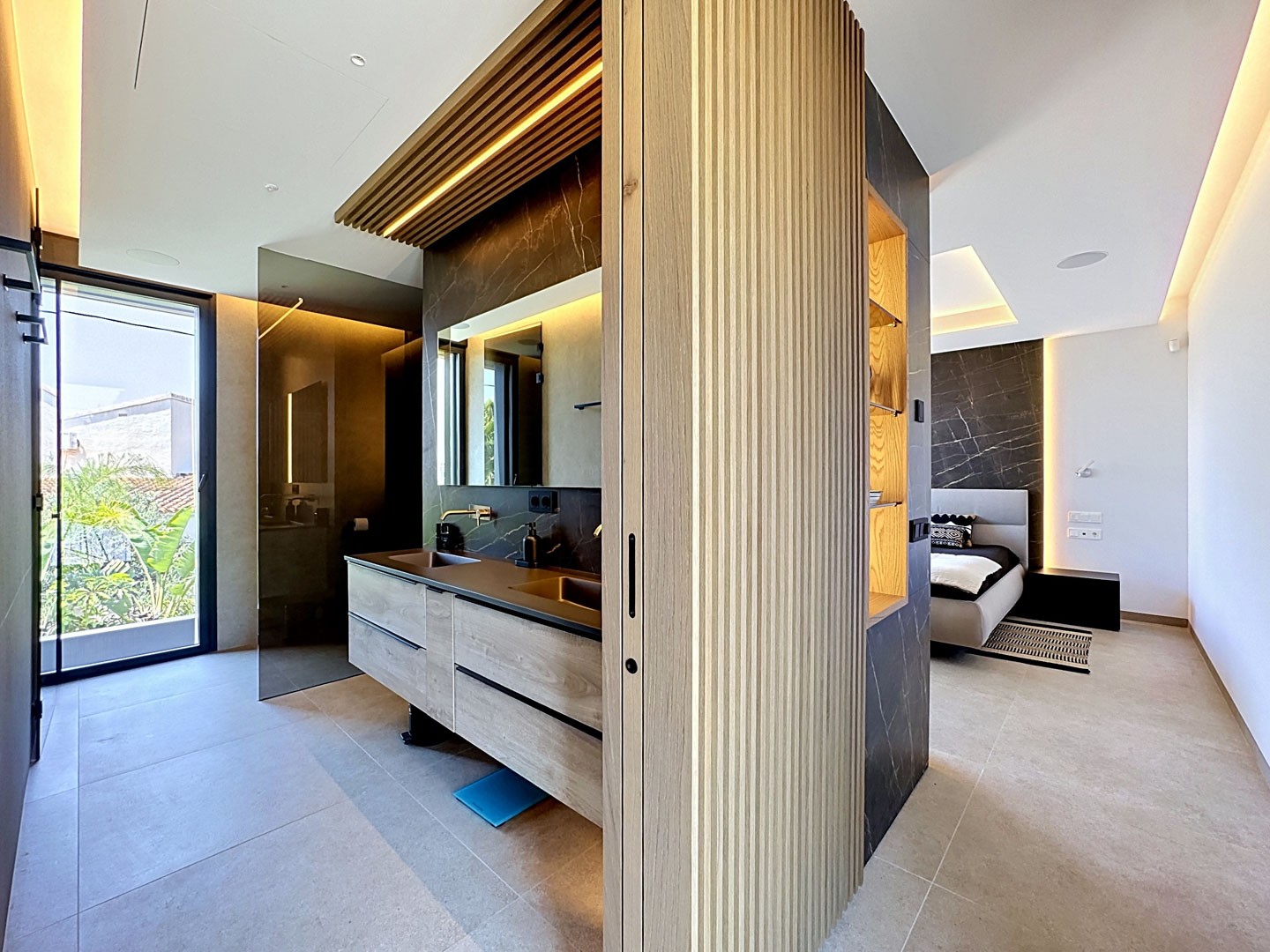 Salle de bain principale intégrée avec double vasque en bois, comptoir en pierre sombre et douche à l'italienne. Une grande porte vitrée s'ouvre sur la verdure extérieure luxuriante.