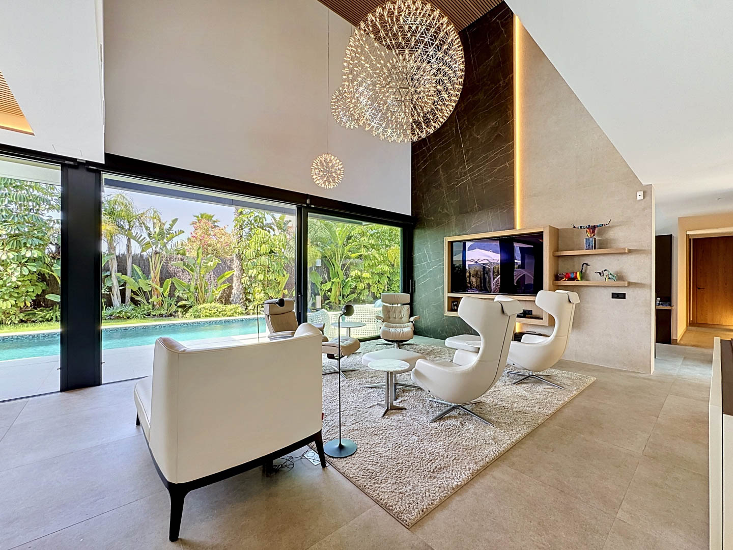 Salon contemporain à double hauteur avec mur d'accent effet marbre foncé, lustres sphériques et grandes portes vitrées donnant sur une piscine privée et un jardin tropical luxuriant.