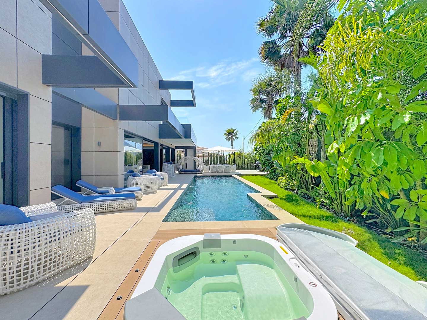 Terrasse extérieure avec piscine rectangulaire et jacuzzi intégré. Le mobilier de salon blanc et bleu s'harmonise avec la façade moderne de la villa et le jardin tropical luxuriant.