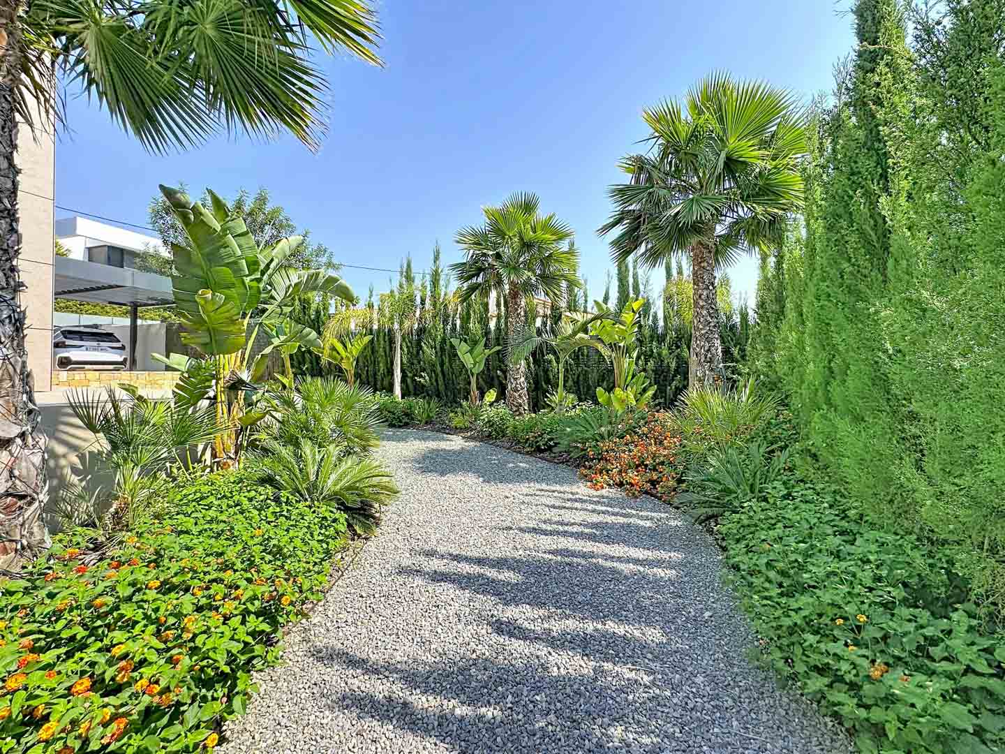 Un chemin de gravier sinueux traverse des jardins tropicaux luxuriants, ornés de palmiers divers, de fleurs colorées et de haies de cyprès, menant à la villa moderne et son abri voiture.