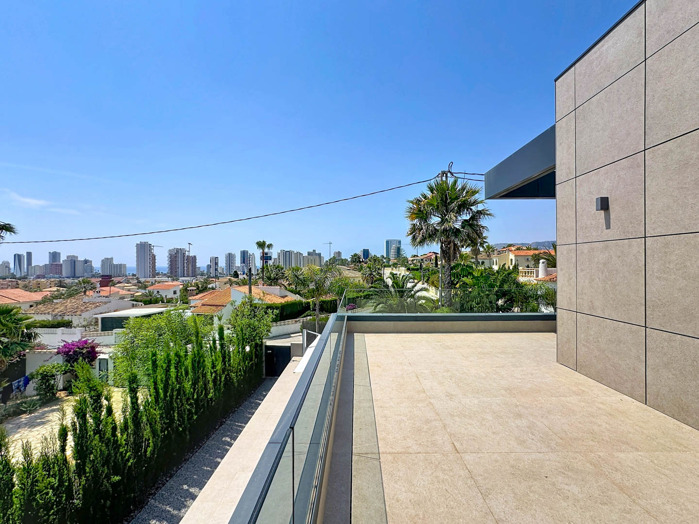 Vaste terrasse supérieure avec carrelage clair et garde-corps en verre, offrant des vues panoramiques sur la skyline de Calpe, la mer et la végétation méditerranéenne. Façade moderne à panneaux sur la droite.