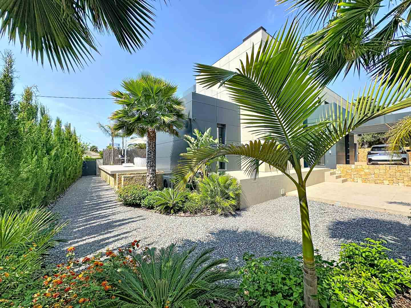 Vue extérieure de la villa avec façade minimaliste grise. Jardin paysager avec allées de gravier, palmiers et végétation tropicale, encadré par une haie haute sous un ciel bleu clair.