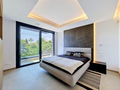 Chambre avec lit design, tête de lit rembourrée et mur d'accent en marbre foncé. De grandes baies vitrées coulissantes donnent sur un balcon avec vue sur la végétation luxuriante, complétées par un éclairage LED intégré.