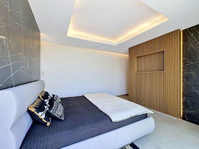 Chambre moderne avec lit tapissé gris clair, panneaux muraux effet marbre foncé et un mur central à lattes de bois avec éclairage intégré et étagère encastrée. Le plafond intègre un éclairage périmétrique.