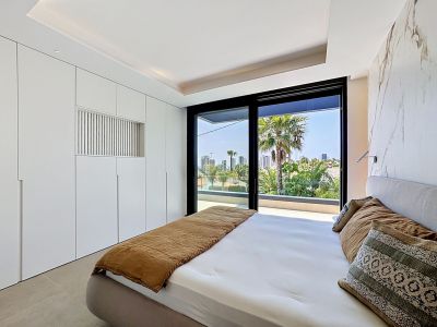 Chambre moderne avec placards intégrés blancs et mur d'accent effet marbre. De grandes portes vitrées s'ouvrent sur une terrasse privée avec vue sur les palmiers et la ville.