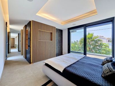 Chambre principale avec un mur d'accent en lattes de bois éclairées et un panneau en marbre foncé. De grandes baies vitrées offrent une vue sur la verdure luxuriante et l'architecture locale.