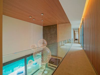 Couloir supérieur avec garde-corps en verre et plafond à lattes de bois, surplombant un salon double hauteur avec lustres sphériques et piscine.