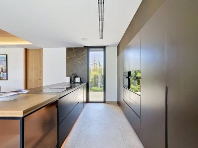 Cuisine design avec meubles sombres sans poignées et électroménager Miele intégré. L'îlot central présente un plan de travail noir, une plaque et un évier, complété par un mur en pierre veinée et un accès jardin.