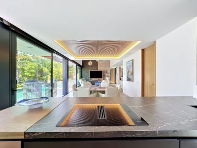Cuisine design avec plan de travail en marbre veiné et plaque Miele intégrée, ouverte sur le salon au plafond en lattes de bois éclairé et accès direct à la piscine.