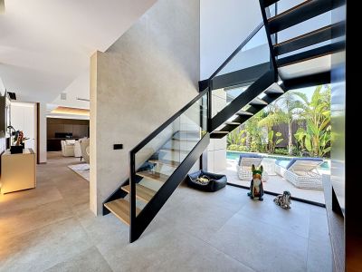 Escalier moderne à marches ouvertes avec garde-corps en verre et métal noir, reliant l'intérieur lumineux et offrant une vue sur la piscine et le jardin tropical.