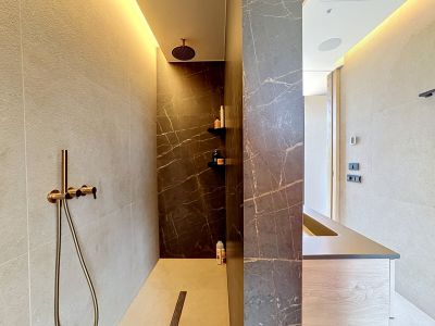 Espace douche contemporain avec carrelage effet marbre foncé veiné et murs clairs texturés. Comprend une douche pluie et douchette en bronze, ainsi qu'un éclairage LED subtil. Une vasque minimaliste est visible.