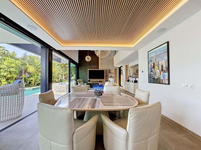 Espace repas élégant avec table ovale et chaises rembourrées, caractérisé par un plafond à lattes de bois éclairé et un accès direct à la piscine et aux jardins tropicaux.