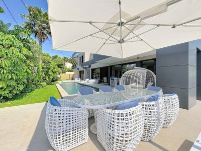 Espace repas extérieur avec table en verre et chaises tressées blanches aux coussins bleus, abrité par un grand parasol. La piscine privée et le jardin tropical bordent la façade moderne de la villa.