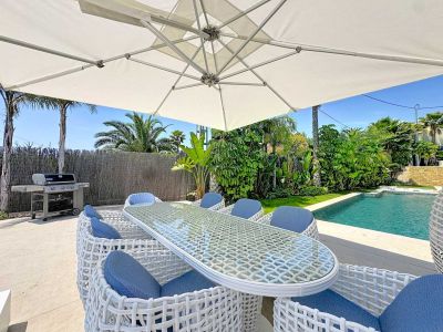 Espace repas extérieur sous un grand parasol blanc, avec table en verre et chaises tressées blanches aux coussins bleus, jouxtant la piscine et un jardin tropical.