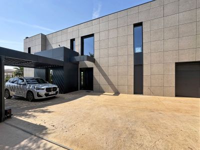 Extérieur contemporain de la villa avec façade en panneaux clairs et entrée anthracite. Un carport à lames protège le véhicule, jouxtant une allée pavée texturée et le garage intégré.