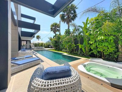L'espace extérieur moderne présente une piscine rectangulaire, un jacuzzi intégré sur une terrasse en bois et un mobilier de salon tressé blanc avec coussins bleus. Des jardins tropicaux luxuriants offrent intimité.