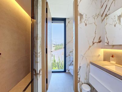 Salle de bain moderne avec douche à l'italienne carrelée de beige et murs effet marbre veiné. Le meuble-lavabo suspendu intègre un éclairage, et une fenêtre toute hauteur offre une vue extérieure.
