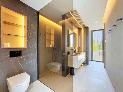 Salle de bain moderne avec murs effet marbre foncé, meuble-lavabo flottant en chêne à double vasque et mur de lattes de bois éclairées. Paroi vitrée pour la douche à l'italienne et toilettes suspendues.