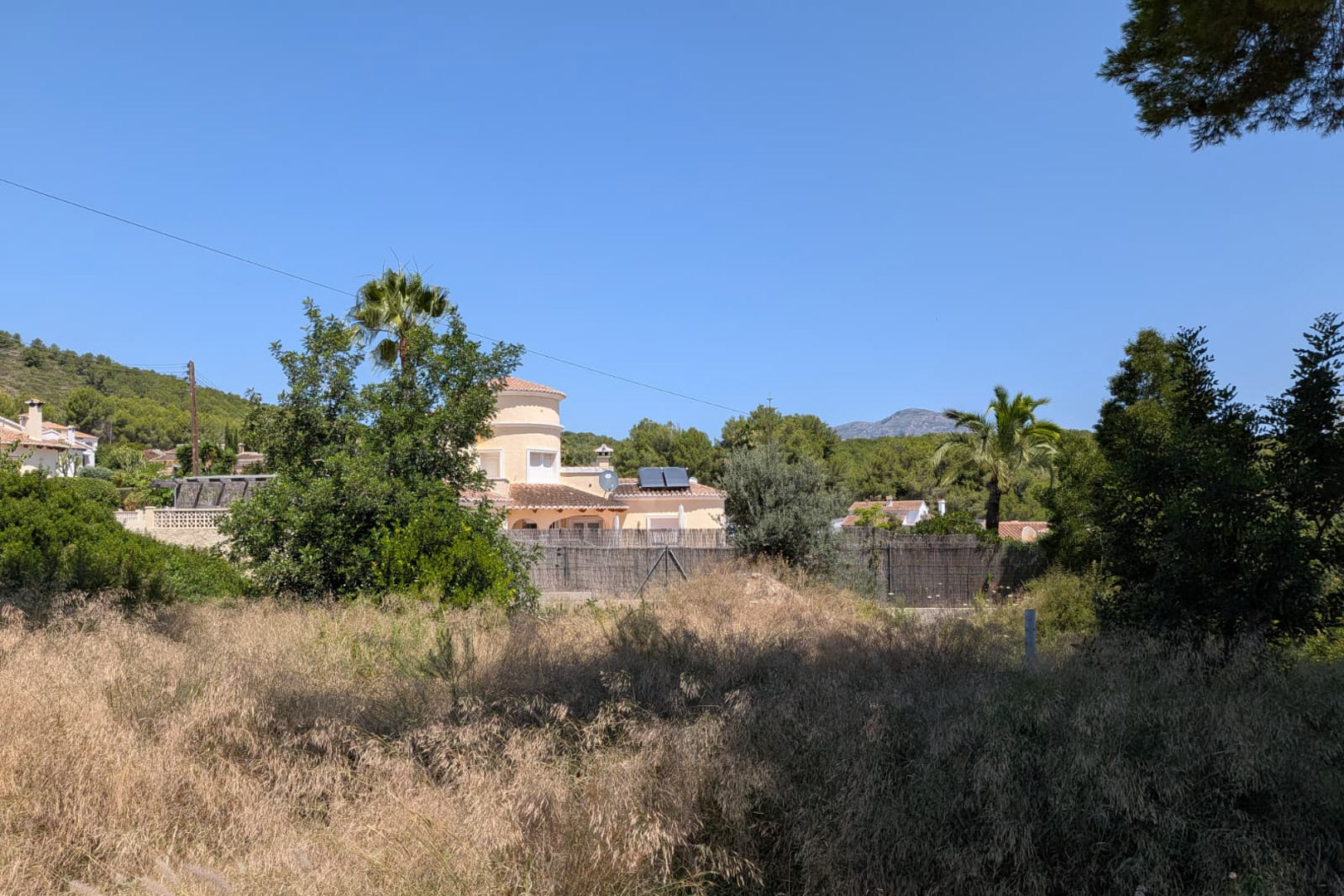 Parcelle avec végétation spontanée et herbe sèche, encadrée par des maisons résidentielles de style méditerranéen aux toits de tuiles et panneaux solaires, et un paysage montagneux sous un ciel clair. Parcelle avec végétation spontanée et herbe sèche, encadrée par des maisons résidentielles de style méditerranéen aux toits de tuiles et panneaux solaires, et un paysage montagneux sous un ciel clair.