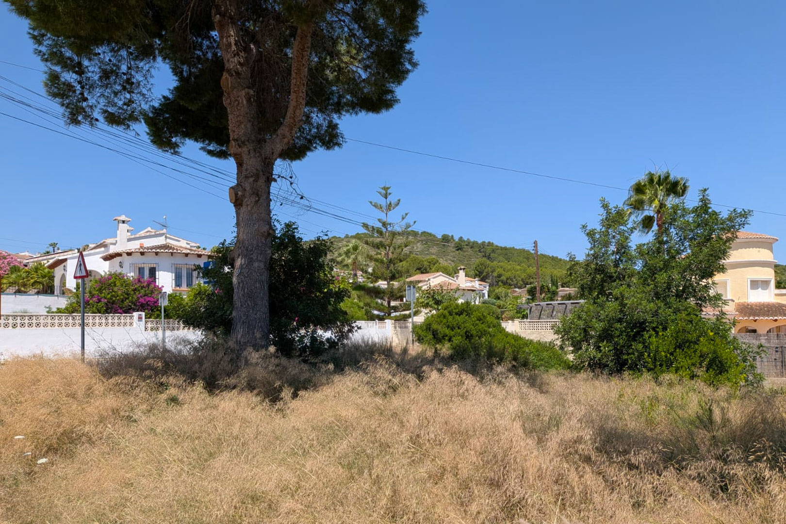 Vaste parcelle de terrain avec végétation naturelle sèche, bordée de villas méditerranéennes et d'une colline boisée sous un ciel bleu clair. Vaste parcelle de terrain avec végétation naturelle sèche, bordée de villas méditerranéennes et d'une colline boisée sous un ciel bleu clair.
