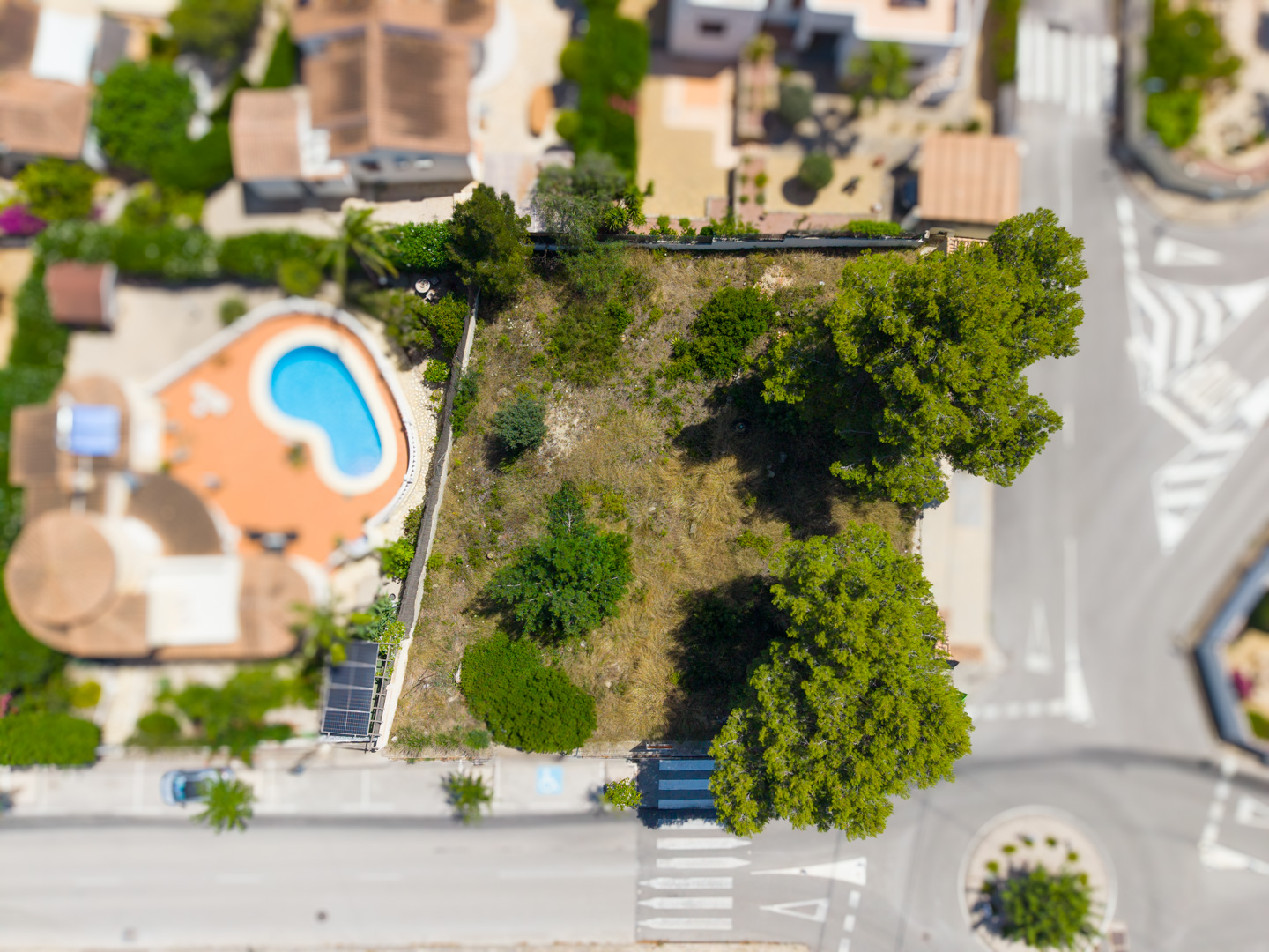 Vue aérienne d'un terrain constructible plat avec végétation clairsemée, bordé par une route résidentielle et une propriété voisine avec piscine. Vue aérienne d'un terrain constructible plat avec végétation clairsemée, bordé par une route résidentielle et une propriété voisine avec piscine.