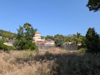 Parcelle avec végétation spontanée et herbe sèche, encadrée par des maisons résidentielles de style méditerranéen aux toits de tuiles et panneaux solaires, et un paysage montagneux sous un ciel clair.