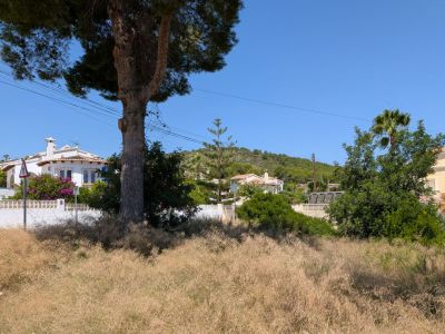 Vaste parcelle de terrain avec végétation naturelle sèche, bordée de villas méditerranéennes et d'une colline boisée sous un ciel bleu clair.