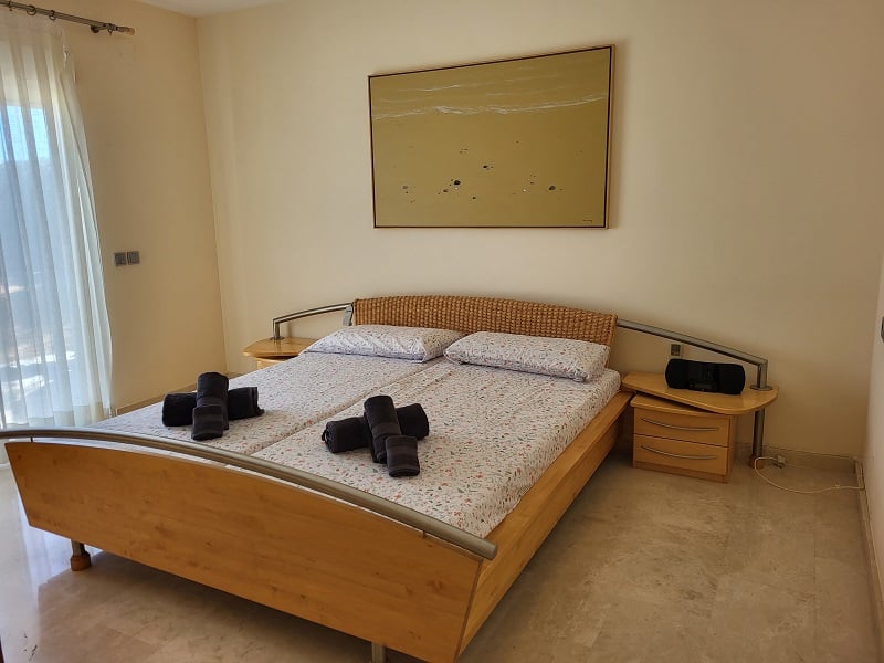 Chambre à coucher avec un lit en bois, une tête de lit tressée et des accents métalliques. Deux tables de chevet avec tiroirs et un tableau abstrait. La lumière naturelle pénètre par une fenêtre avec des rideaux voilés.