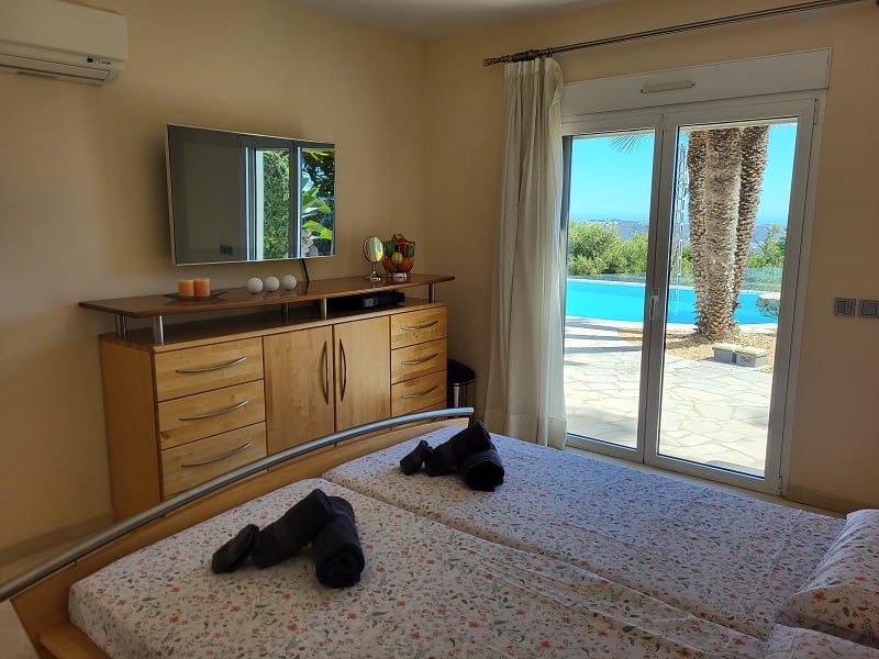 Chambre avec lits jumeaux, télévision murale au-dessus d'une commode en bois et accès direct à la terrasse extérieure avec piscine et palmiers.
