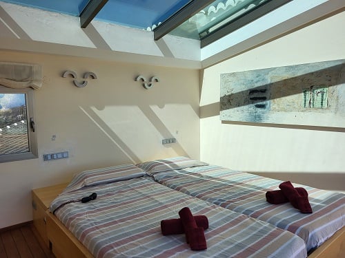 Chambre lumineuse avec deux lits simples, parures de lit rayées et serviettes rouges. Un puits de lumière zénithal éclaire la pièce, agrémenté d'un tableau abstrait et d'une fenêtre.