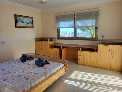 Chambre lumineuse avec lit en bois clair, bureau intégré et rangements. Les fenêtres offrent des vues panoramiques sur la côte, inondant la pièce de lumière naturelle.