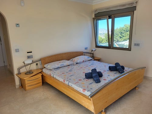 Chambre lumineuse avec lit en bois clair, tête de lit en osier et table de chevet. La fenêtre ouverte encadre des vues sur la verdure et les montagnes.